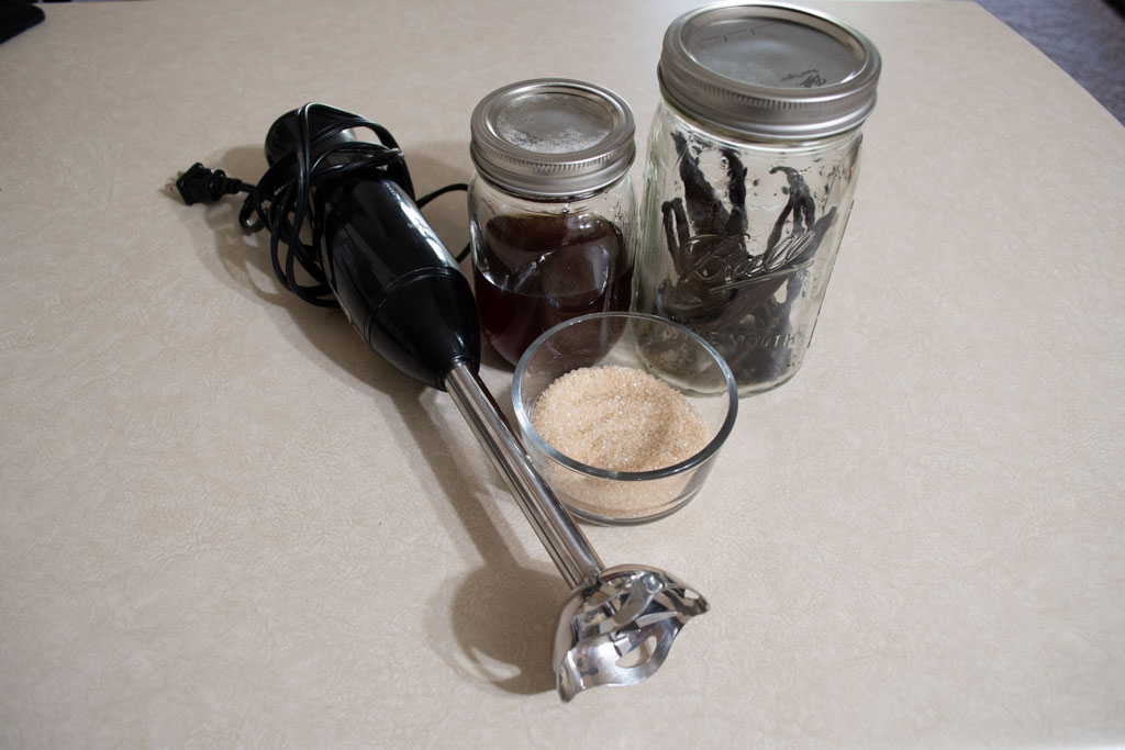 Collected ingredients to make vanilla bean paste including an immersion blender, a glass bowl of cane sugar, one pint jar with vanilla extract and one quart jar with vanilla beans.